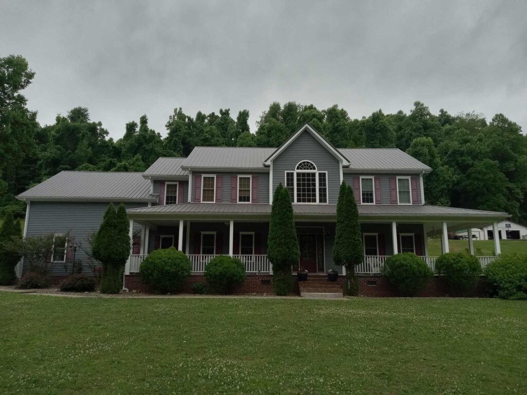 A large, two-story house with gray siding and a gabled roof sits in front of dense green trees. The front porch is surrounded by tall bushes. The sky is overcast.