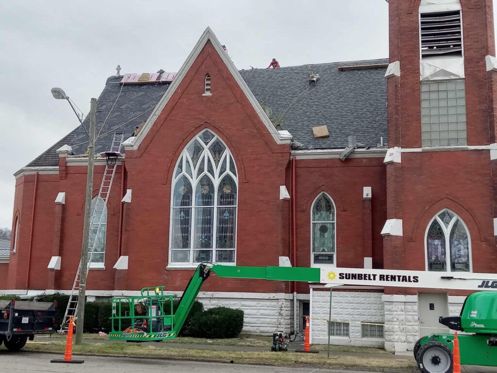 A red brick church with large arched stained glass windows is under maintenance. Ladders and a green lift from Sunbelt Rentals are in use. Overcast skies suggest a calm day.