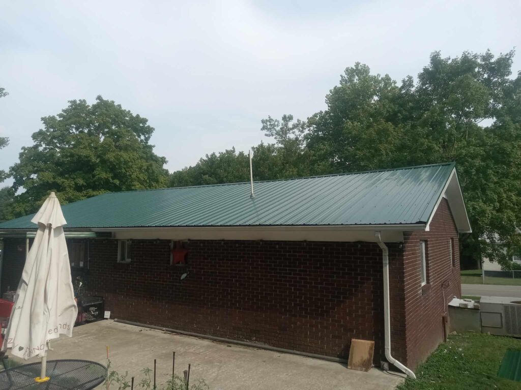 A single-story brick house with a green metal roof and a red chimney is pictured. In the foreground, a closed patio umbrella stands on a concrete patio. Lush green trees surround the house under a cloudy sky.