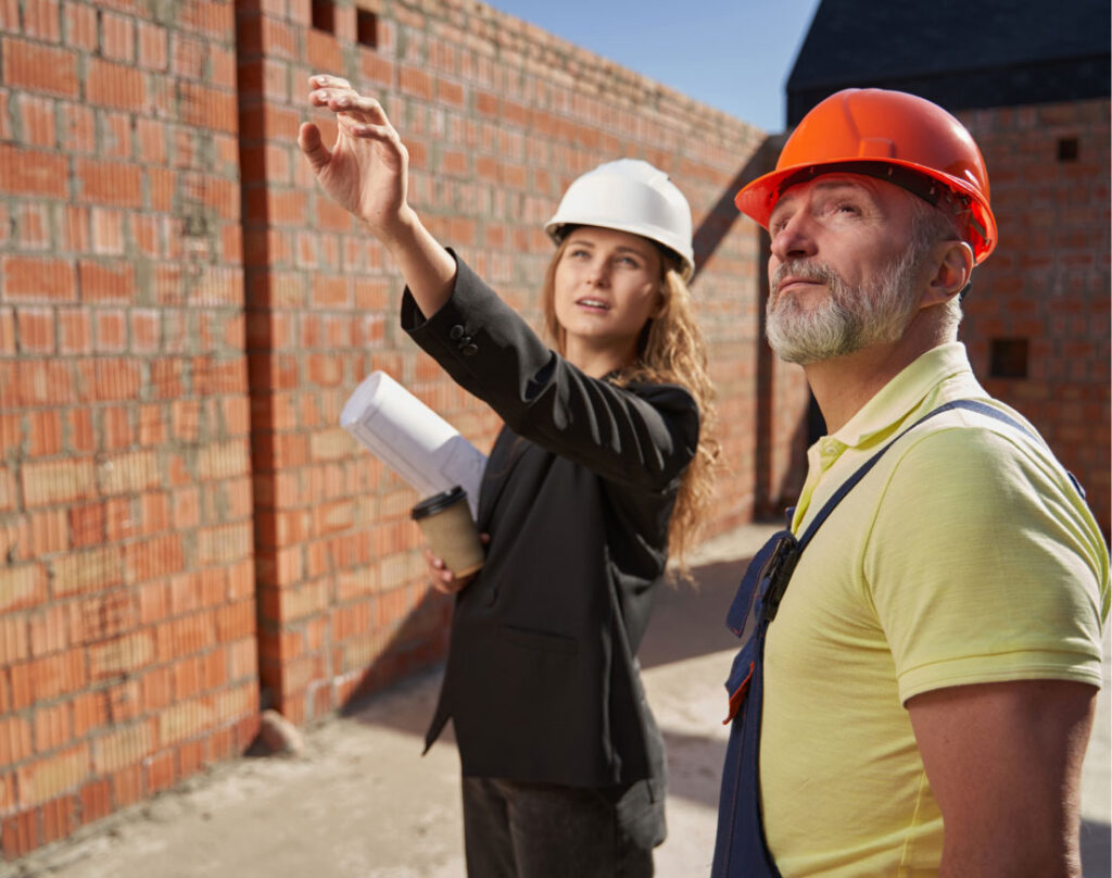A woman in a black blazer and white hard hat gestures towards a brick wall, holding blueprints and coffee. A man in a yellow shirt and orange hard hat looks on, conveying teamwork and focus at a construction site.